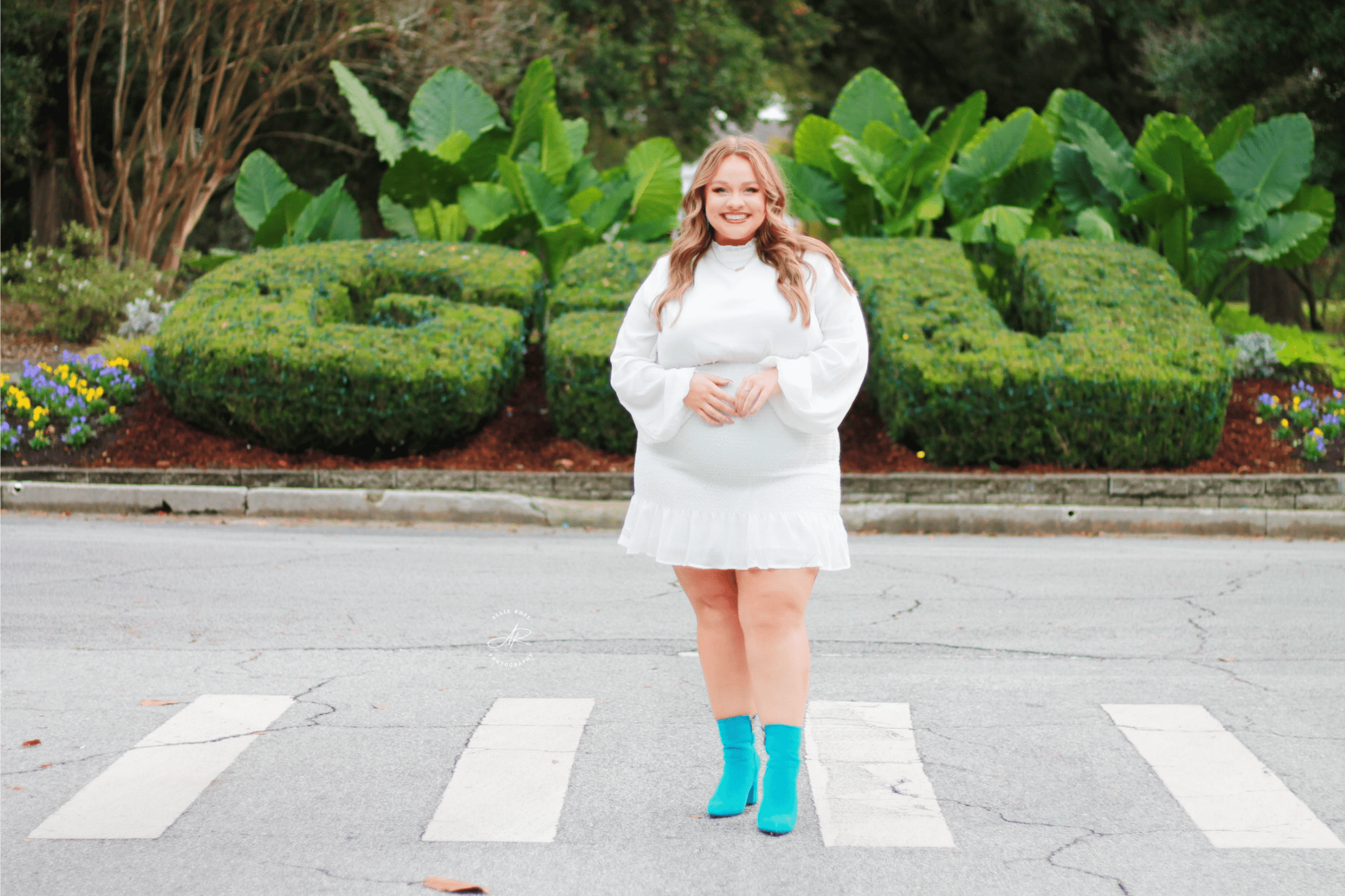 Gracie McMillan stands in a white dress and teal boots in front of the GSU bushes. Photo Credit: Allie Rose Photography