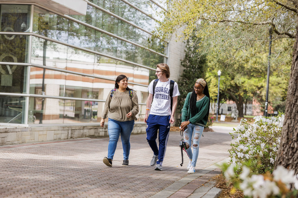 Three college students walk together on a tree-lined campus pathway. Two women and one man are smiling and talking, carrying backpacks and books, with a modern glass building in the background.