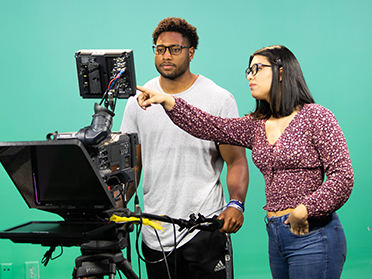 Two people stand in front of a green screen. One person points at a monitor attached to a camera while the other watches, both appearing focused on the screen.