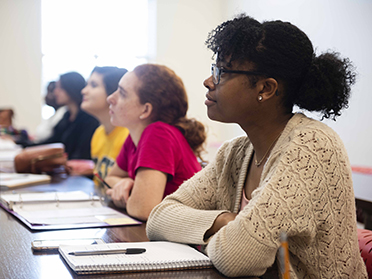 A group of students sits at desks in a classroom, attentively listening and taking notes. The focus is on a student in the foreground wearing glasses and a beige sweater.