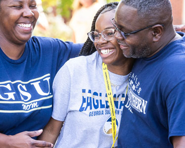 Three people smile and hug each other outdoors. The young woman in the center wears glasses and a Georgia Southern shirt, while the two adults on either side wear GSU and Georgia Southern shirts.