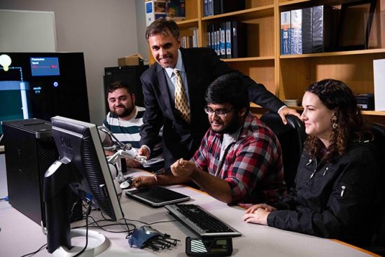Three students learning computer tech from a professional in a classroom setting.