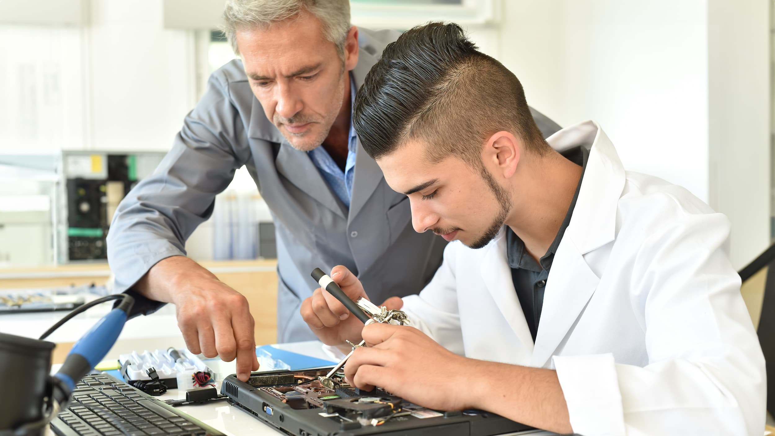 Student in lab coat studying a circuit board with professor to learn electrical and computer engineering.