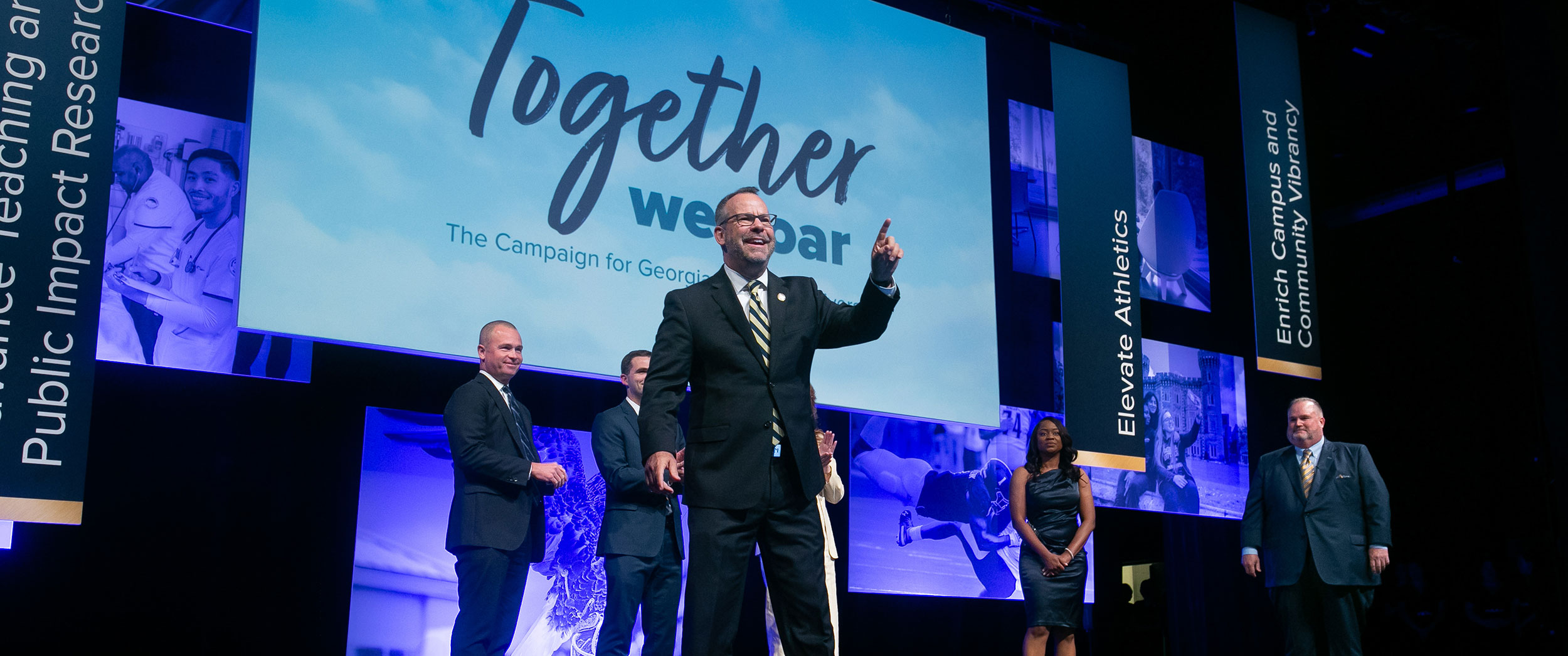 A dynamic stage event photograph showing a President Kyle in a suit gesturing excitedly to the audience in front of large digital screens displaying the words 