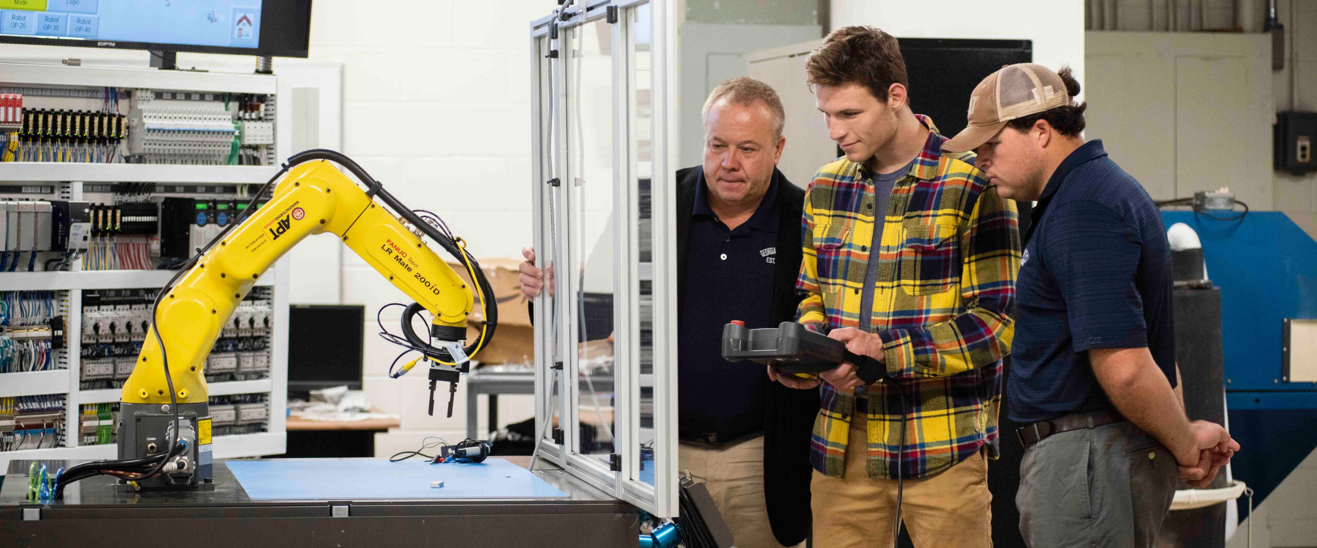A male student in a yellow plaid flannel shirt operating a handheld controller for a yellow FANUC robotic arm in a robotics lab while two men look on.