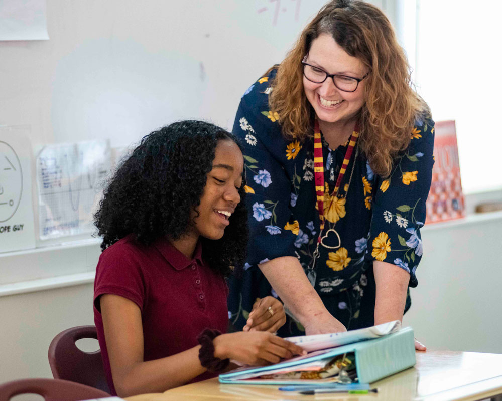 A student sitting at a desk smiles while looking at her open book, as a teacher standing beside her leans in and smiles, engaging warmly in conversation.