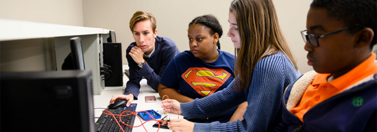 Three diverse students looking focused while working together at a computer screen in a classroom or lab.