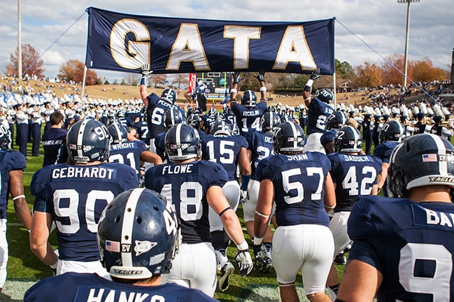 Football players in navy uniforms run onto the field toward a large banner reading GATA, with a marching band and a crowd in the background. The scene is energetic and captures the excitement before a game.