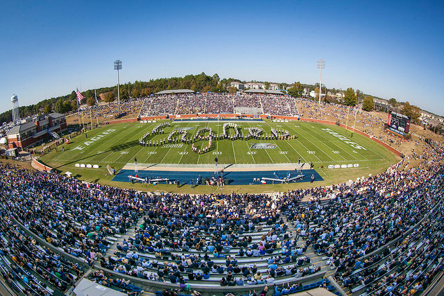 A panoramic view of a packed football stadium during a game, with a marching band on the field spelling out Eagles in formation, surrounded by cheering fans in the stands.