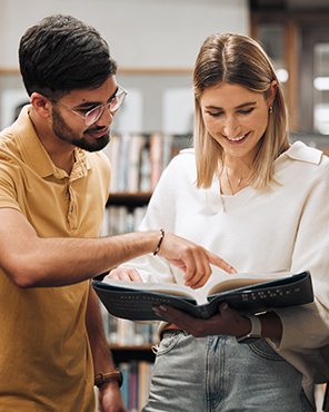 A male student wearing glasses and a yellow polo shirt pointing at a page in a book held open by a smiling female student in a library setting.