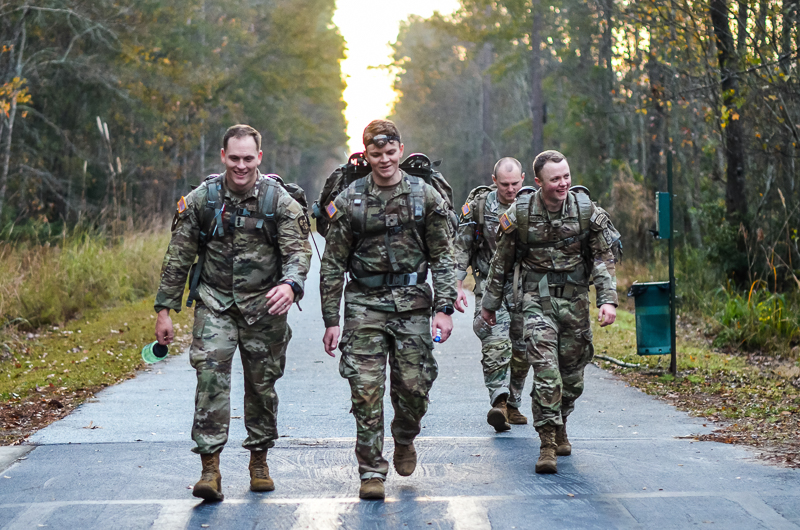 image_f3a3a1.jpg: A group of four smiling Army ROTC cadets in full combat gear with rucksacks walking together down a paved woodland trail during a road march.