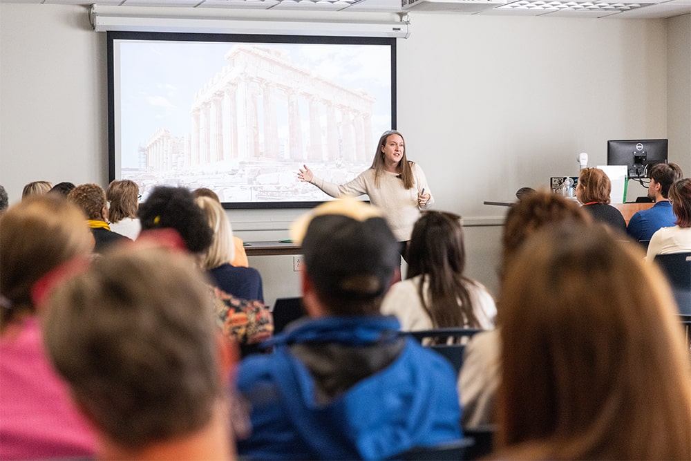 A person is giving a presentation to a seated audience in a classroom. The presenter gestures toward a projected image of ancient ruins, resembling a Greek architectural structure. The audience sits attentively, with some taking notes.