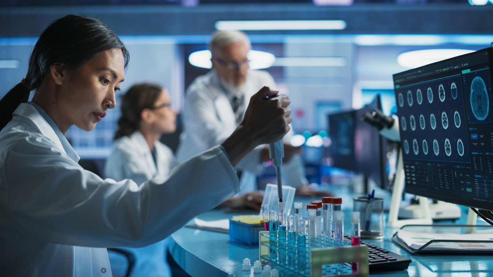 A modern laboratory setting showing a female scientist in a white lab coat using a pipette to work with liquid samples in test tubes, while a computer monitor nearby displays rows of brain scan/MRI imagery and other researchers collaborate in the background.