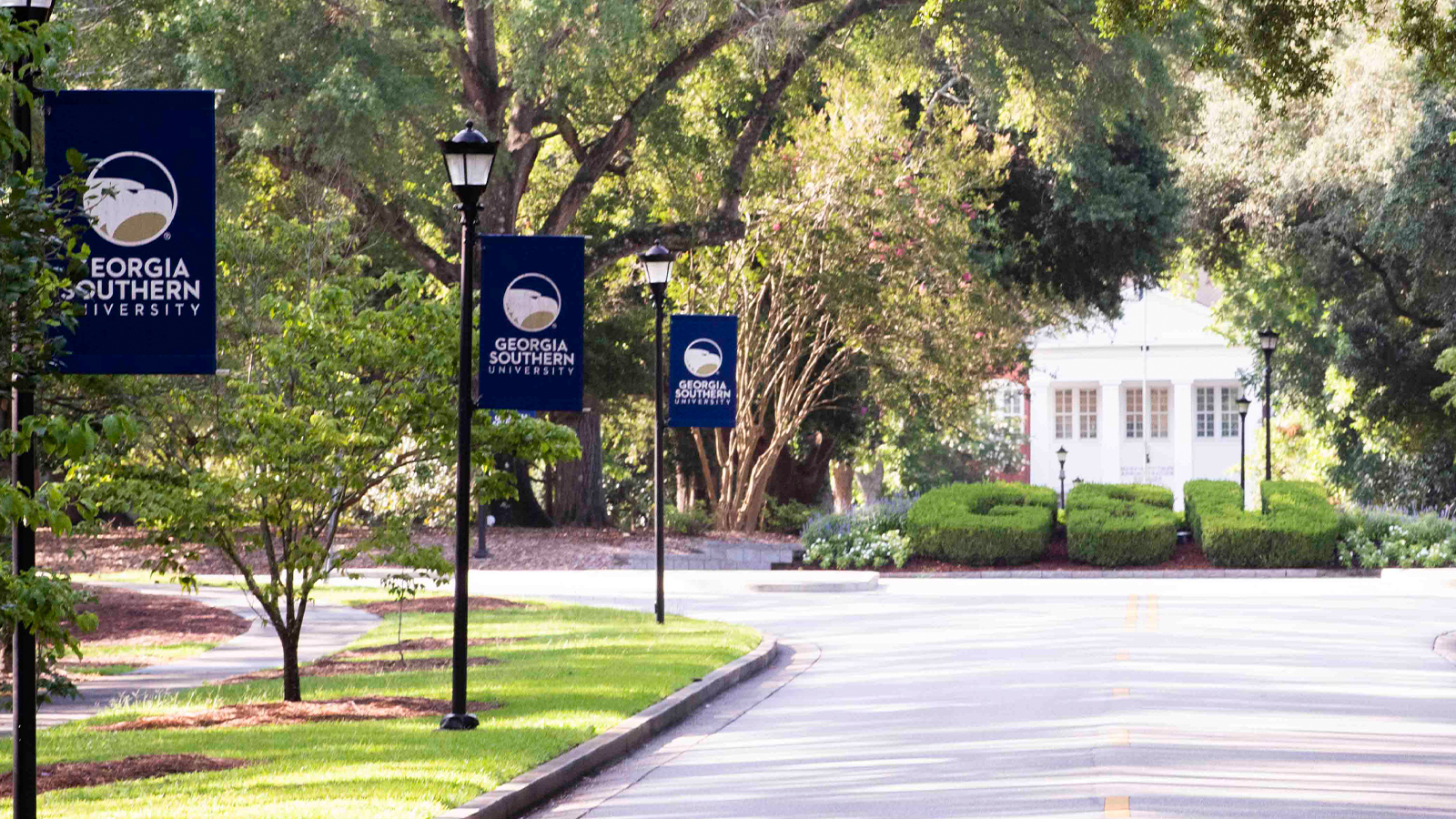 A scenic, sunlit view looking down a campus roadway lined with lamp posts flying dark blue Georgia Southern University banners. In the background, large hedges are meticulously shaped into the letters "GSU" in front of a white building nestled among lush green trees.