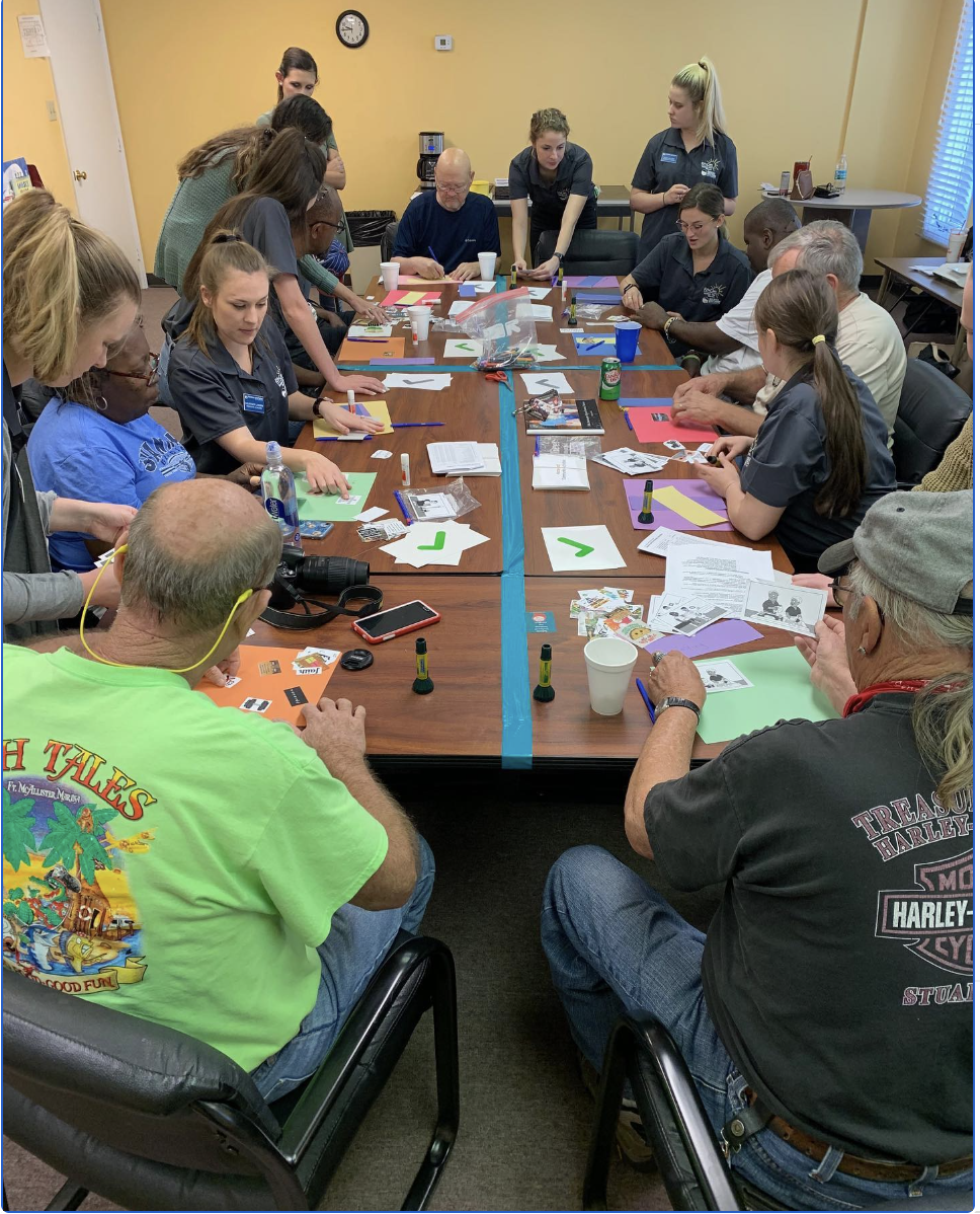 In a brightly lit room, a diverse group of people sits around a long wooden table divided by blue tape, actively engaged in a hands-on workshop with colorful paper, glue sticks, and printed materials.