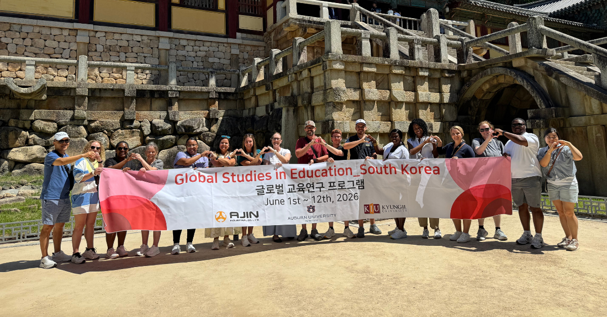 A group of people hold a banner that says "Global Studies in Education_South Korea" which is accompanied by Korean text. They stand in front of a beautiful building making "heart hands" with each other.