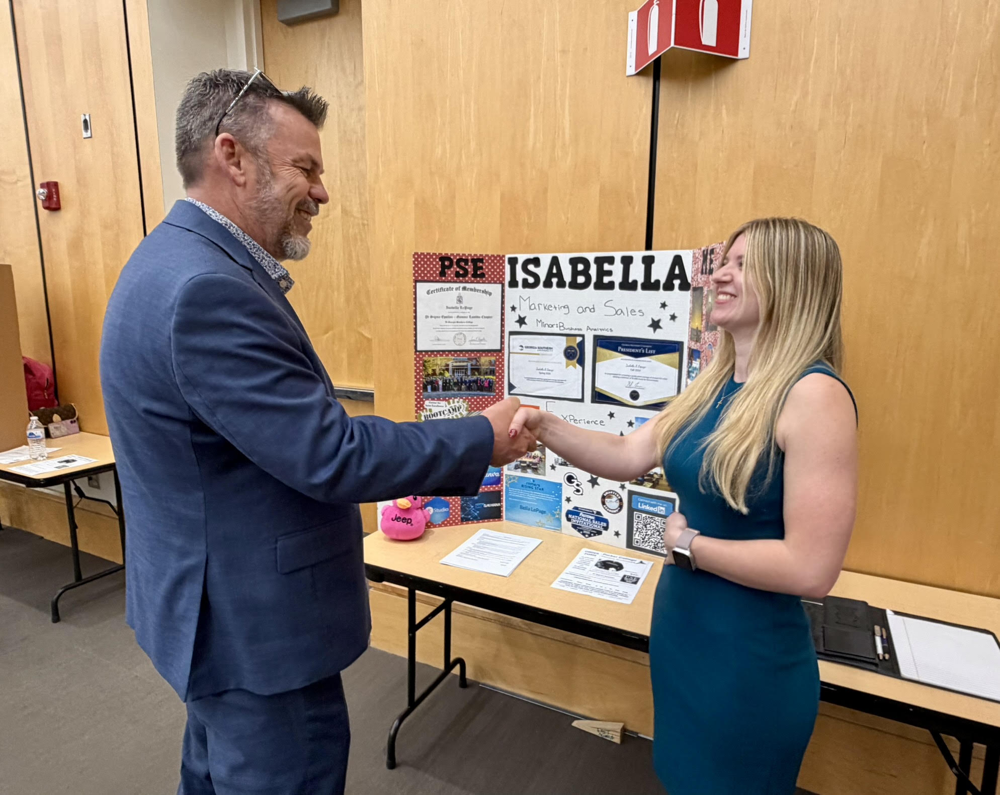 A professional man in a blue suit shaking hands with a smiling female student wearing a teal dress at a reverse career fair. Behind them, a trifold presentation board labeled Isabella highlights her background in marketing, sales, and business analytics, alongside certificates and photos.