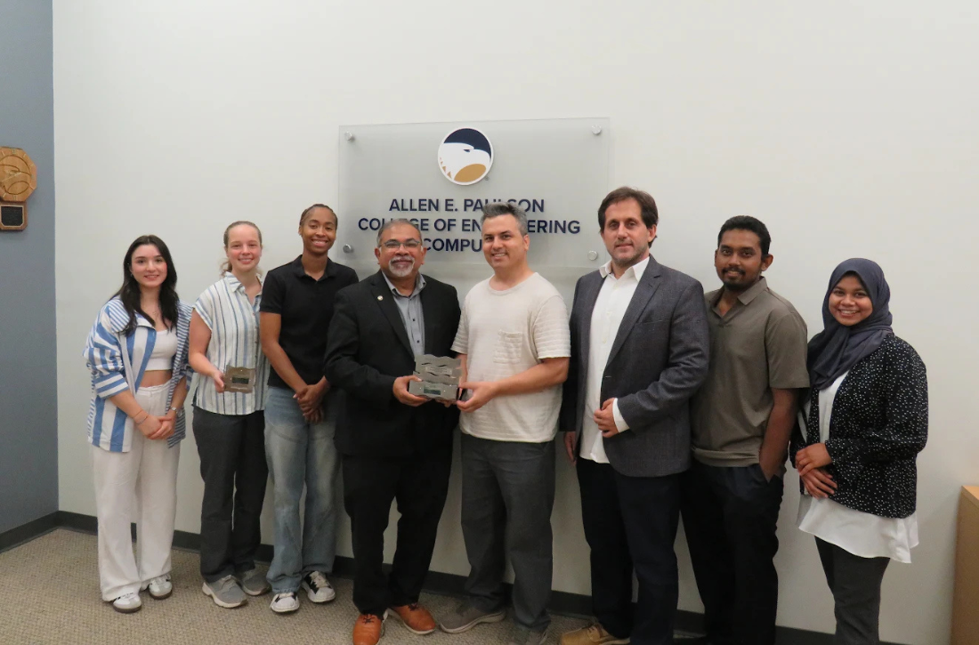 A group of engineering professionals stand in front of a sign that says Allen E. Paulson College of Engineering and Computing holding a square-shaped award and smiling.